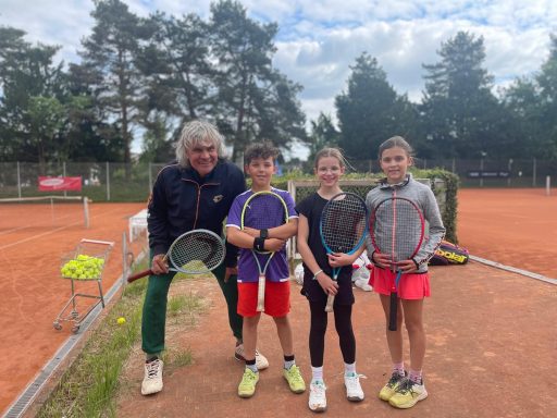 Vier Kinder stehen mit Schlägern auf einem Tennisplatz, ein Trainer lächelt daneben.