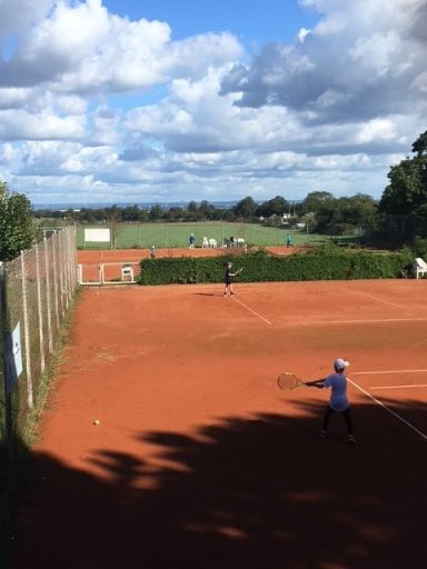Tennisplatz mit zwei Spielern, einem im Vordergrund und einem im Hintergrund. Wolkiger Himmel.