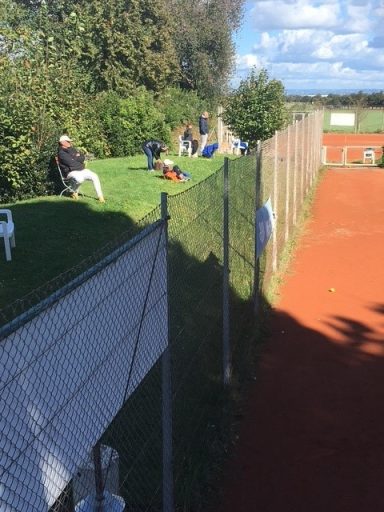 Tennisplatz mit Zuschauern, Bäumen im Hintergrund und blauer Himmel.