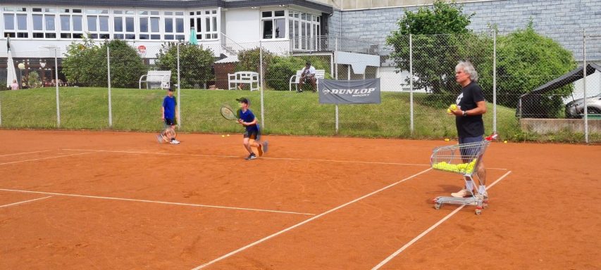 Zwei Kinder spielen Tennis auf einem Sandplatz, ein Erwachsener steht am Rand.